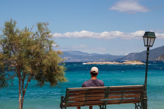 Мужчина смотрит на Средиземное море и горы, сидя на скамейке возле дерева и уличного фонаря