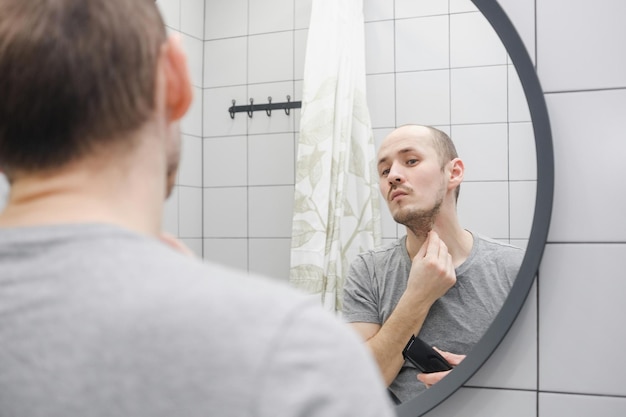 A man looking at mirror in bathroom preparing cutting beard\
using electric razor