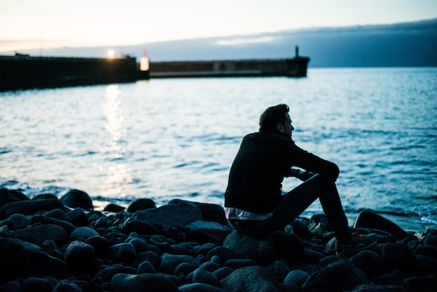 Foto uomo che guarda il mare seduto su una roccia