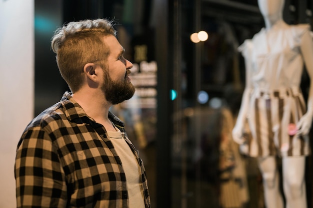 Uomo che guarda la vetrina nel negozio di strada serale e il concetto di consumismo maniacale dello shopping