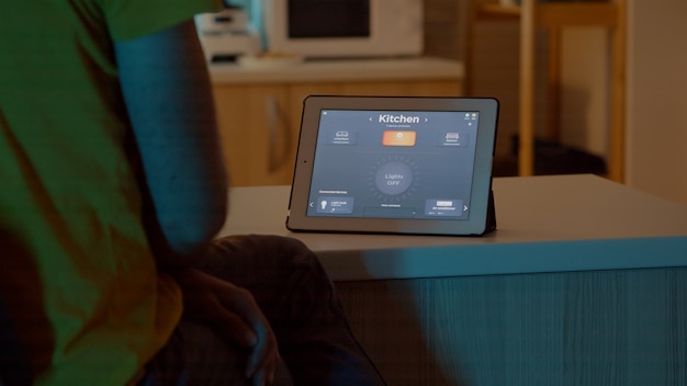 Man looking at tablet in house with automation lighting system, sitting in kitchen switching on bulbs using voice command