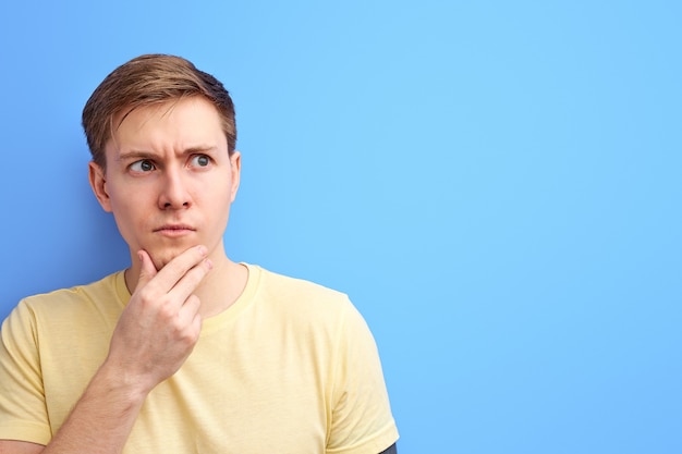 Man looks away and thinks with a serious face, mindful male holding the chin with hand, stand in contemplation isolated over blue background