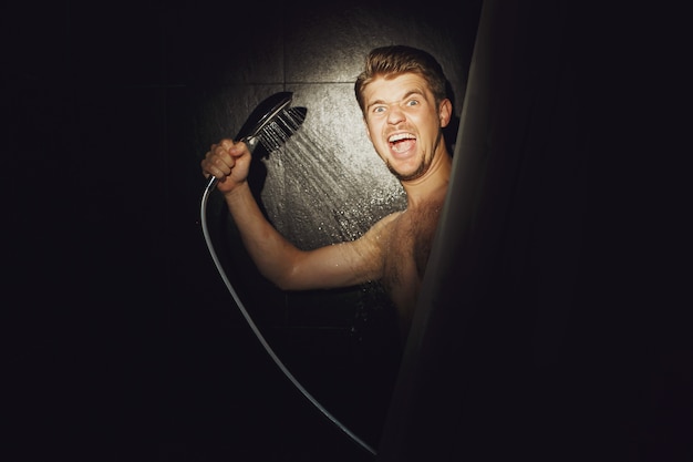 Man looks funny holding shower over him in black bathroom