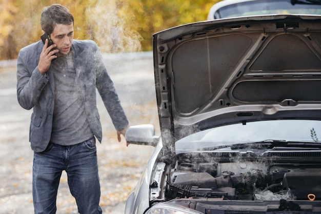 A man looks under the open hood of a car The car broke down on the road The engine is smoking