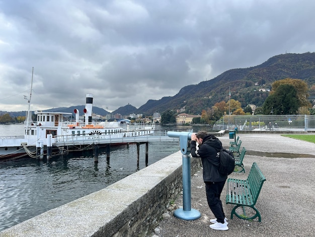 Man looks through a telescope at the sea standing on the shore at the foot of the mountains