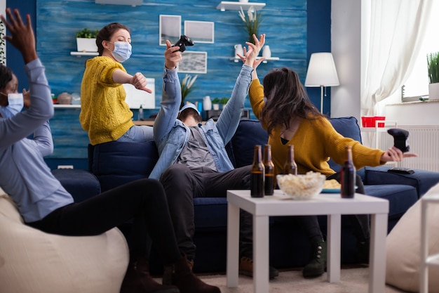 Man losing video game competiton against multi ethnic friends in home living room wearing face mask keeping social distancing as prevention during covid outbreak. new normal party