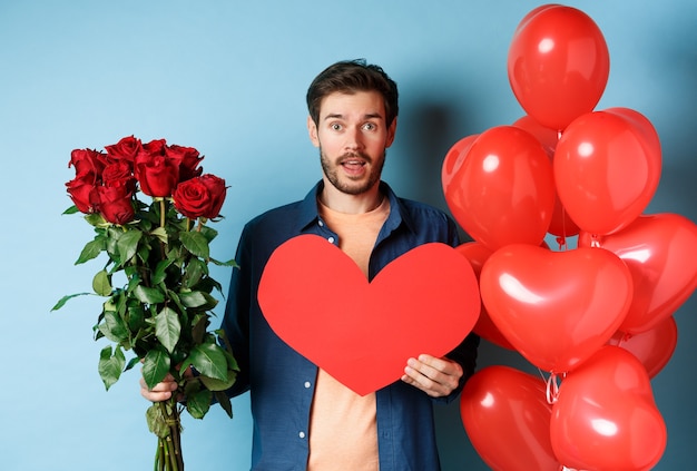 L'uomo innamorato porta regali a sorpresa in un appuntamento romantico, tenendo in mano il mazzo di rose rosse e cuore rosso di san valentino, in piedi vicino a palloncini e guardando l'amante, sfondo blu.