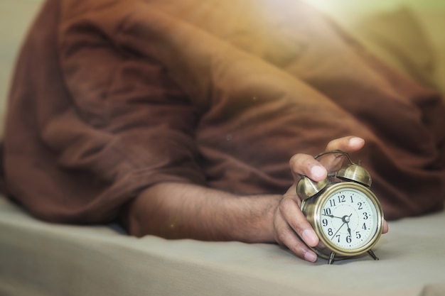 A man lying under a brown blanket is going off the alarm clock with drowsiness.