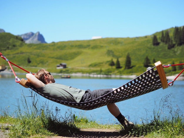 Man lying in hammock at lakeshore against mountains