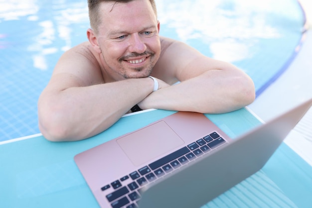 Man lying on side of swimming pool and looking at laptop screen