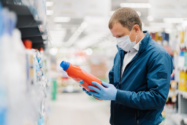 Man makes shopping in supermarket, holds bottle of washing detergent, reads product information, wears protective medical mask and gloves during coronavirus outbreak, chooses something for household