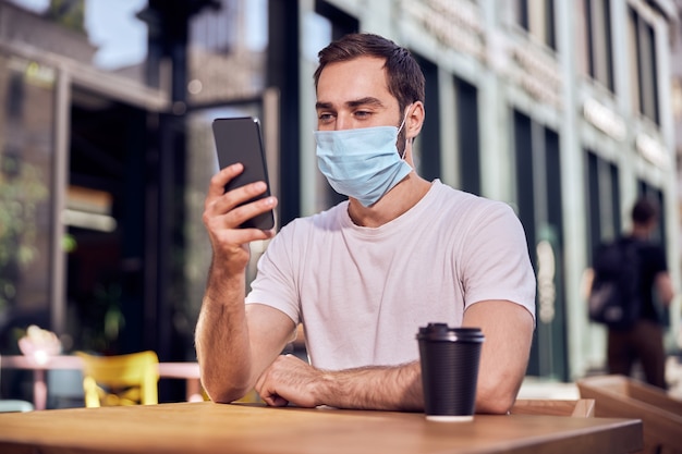 Man in mask is sittiing  with smartphone and coffee