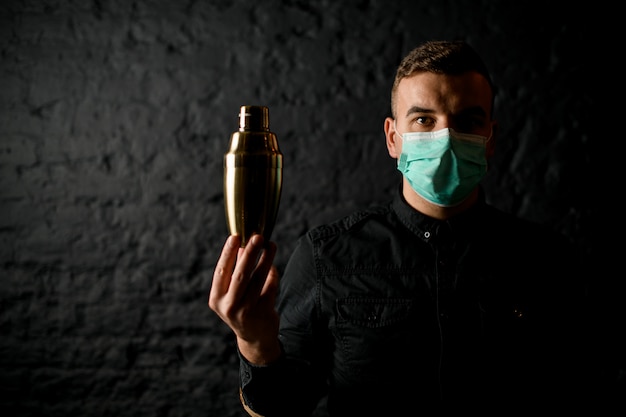 Man in mask stands in dark bar and holds steel shaker in his hand