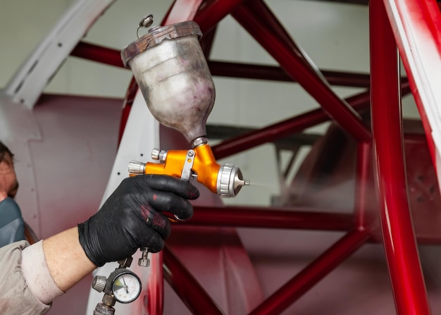 A man master in protective overalls and a mask holds a spray bottle in his hand and sprays red paint onto the frame of the car body after an accident during a repair in a vehicle restoration workshop