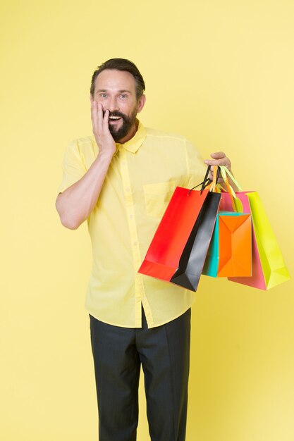 Man mature bearded cheerful face holds shopping bags. Man got unexpectable gifts. Guy surprised by gift yellow background. Presents make our lives more interesting. Bonus for client always good idea.