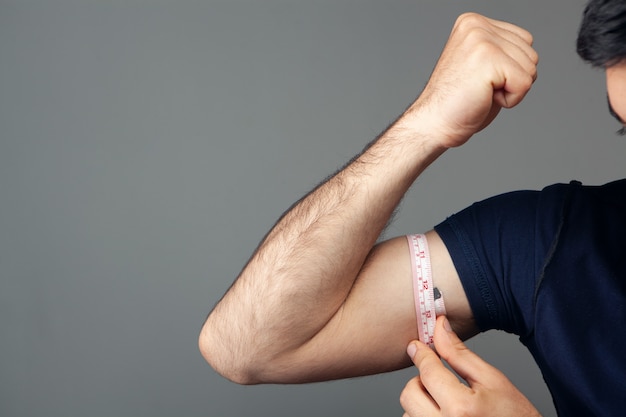 A man measures the girth of his arm with a meter on gray