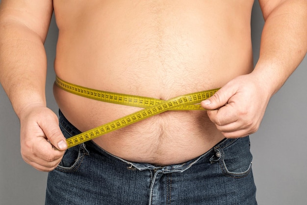 A man measures his fat belly with a measuring tape on a gray background