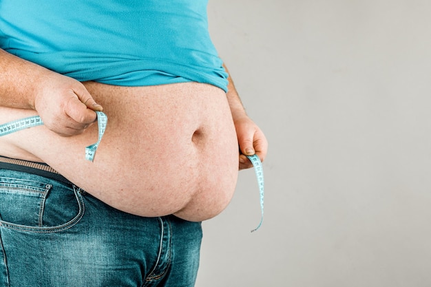 A man measures his fat belly with a measuring tape on a gray background