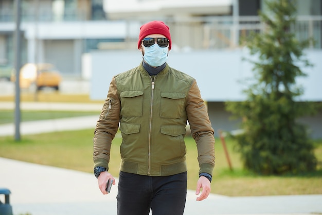 Человек в медицинской маске, чтобы избежать распространения коронавируса, держит свой смартфон на уютной улице.