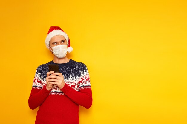 Foto uomo in maschera medica maglione natalizio il cappello di babbo natale tiene in mano lo smartphone e scrive un messaggio