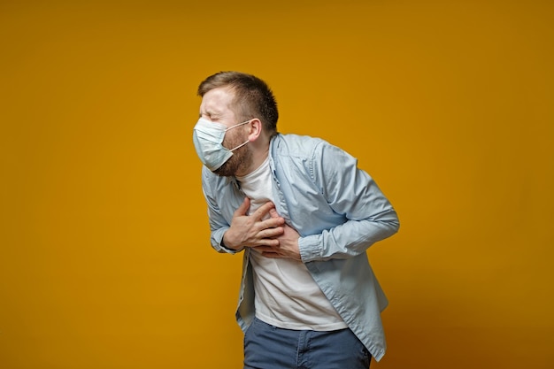 Man in a medical mask coughs holds hands on chest experiencing pain and closes eyes from suffering