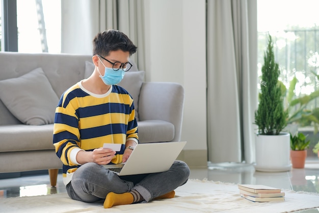 Photo man in medical mask looking at credit card while using laptop near headphones of floor in living room