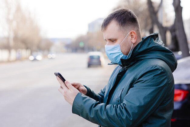 a man in a medical mask on the side of the road with a phone in his hand is calling a taxi