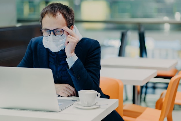 Man in medical mask and spectacles sits in front of modern laptop computer poses in cozy cafeteria reads news about coronavirus concerned about health drinks coffee Epidemic quarantine