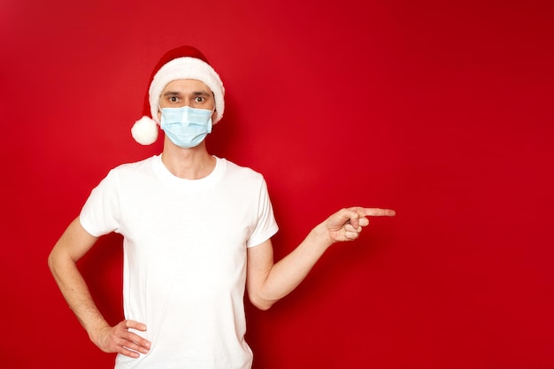 산타클로스 크리스마스 모자를 쓴 의료용 마스크를 쓴 남자는 빈 좌석을 가리킵니다. 격리된 빨간색 스튜디오 배경