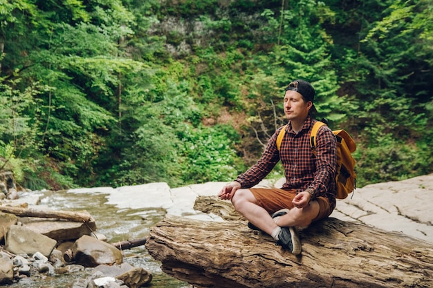 человек в медитативной позе сидит на стволе дерева на фоне леса и реки