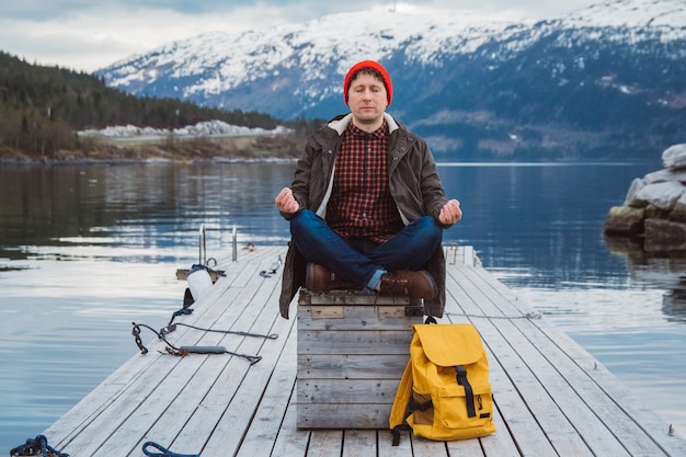 man in a meditative position sitting on a wooden pier on the background of a mountain and a lake