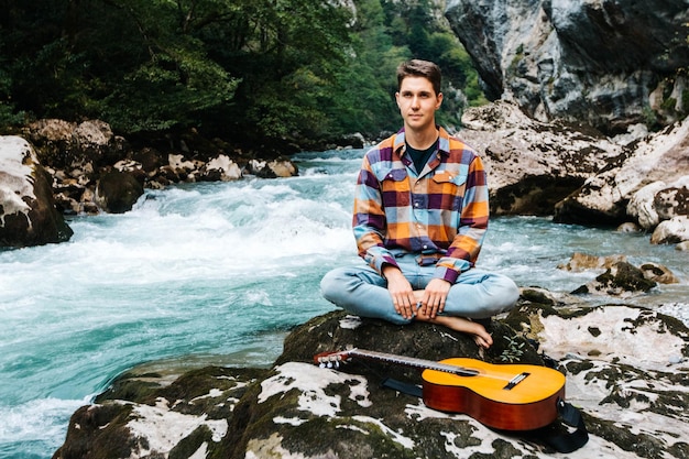 Man in a meditative position with guitar sitting on the bank of a mountain river