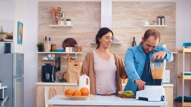 Man mengen van fruit voor smoothie met blender in de keuken. Vrolijk jong stel. Vrolijke familie die samen biologisch gezond vers voedzaam smakelijk sap maakt voor het ontbijt van vers fruit terwijl ze op vakantie zijn