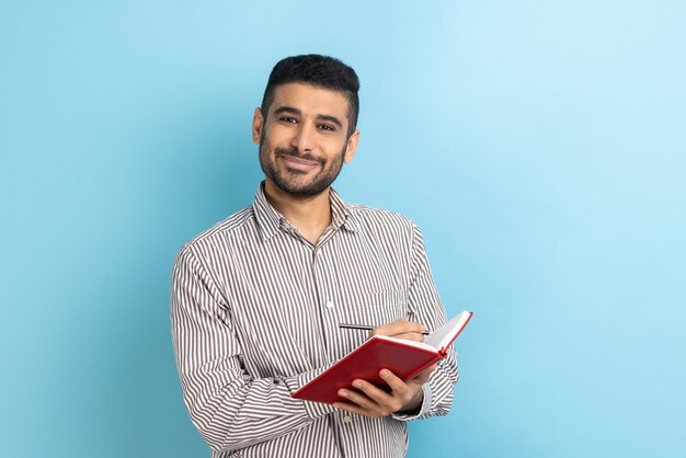 Man met baard die een takenlijst opschrijft en aantekeningen maakt met een pen in een notitieboekje die naar de camera kijkt