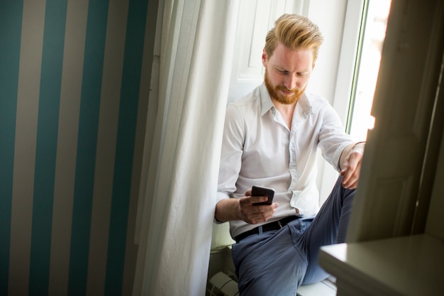 Man met baard en rode haren met behulp van smartphone voor antwoord op berichten en zittend op venster