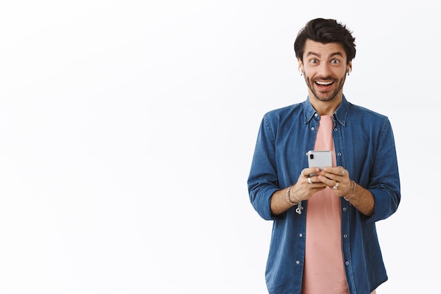 man met baard en stijlvol rommelig kapsel, smartphone vasthoudend, opgewonden en geamuseerde camera grijnzend, goed nieuws ontvangen