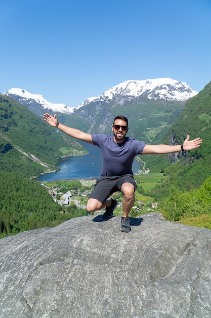 Man met baard en zonnebril die zijn armen op een rots steekt met de Geirangerfjord op de achtergrond en de besneeuwde bergen op een zonnige dag