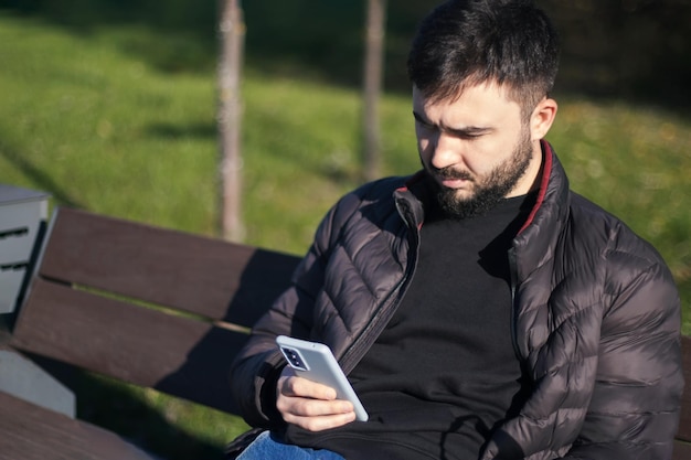 Man met baard zit op de bank in het park. Hij scrolt door het nieuws op zijn smartphone