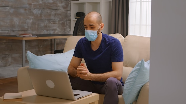 Man met beschermingsmasker tijdens een telefonische vergadering op laptop tijdens coronavirusvergrendeling.