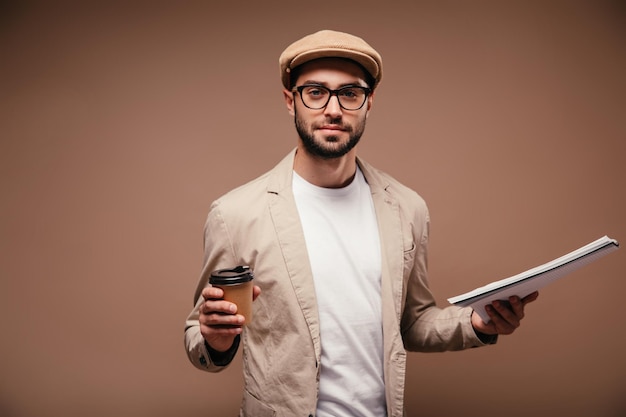 Man met bril en beige pet kijkt in camera met notitieboekje en glas koffie