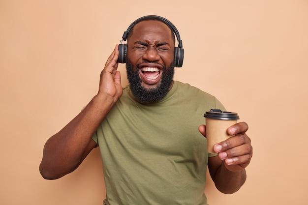 man met dikke baard voelt zich energiek luistert naar audiotrack via koptelefoon houdt papieren kopje koffie geniet van vrije tijd casual gekleed in beige