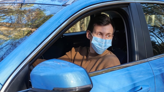 Man met donker haar met medisch masker zit in de auto langs de weg in de buurt van het bos van dichtbij