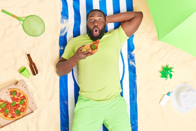 man met donkere huid staart onder de indruk van camera ligt aan zandstrand eet heerlijke pizza gekleed in groen t-shirt en korte broek geniet van ontspanningstijd luie dag