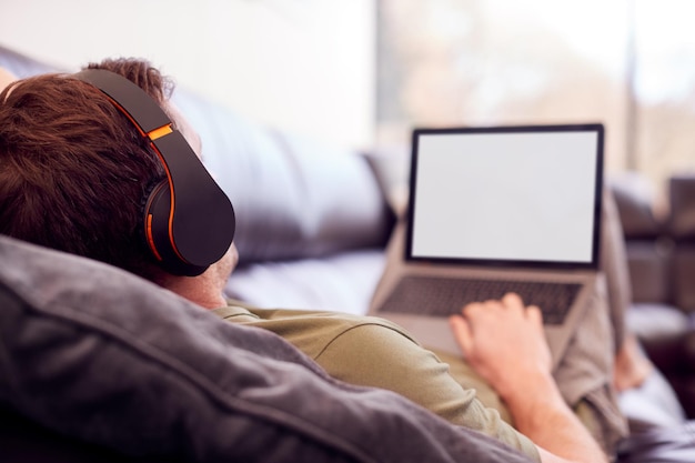 Foto man met draadloze koptelefoon luisteren naar muziek stomen van laptop op de bank thuis in pyjama