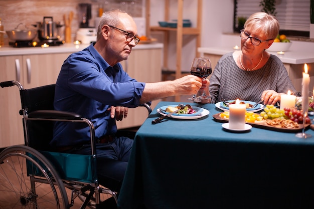 Man met een handicap aan het dineren met vrouw en rammelend wijnglas. Rolstoel geïmmobiliseerd verlamde gehandicapte man dineren met vrouw thuis, genietend van de maaltijd