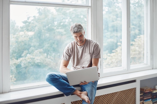 Man met een laptop zittend op de vensterbank in de woonkamer