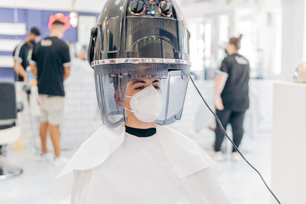 Man met een masker zittend in een kapsalon met een elektrische haardroger om zijn hoofd gewikkeld