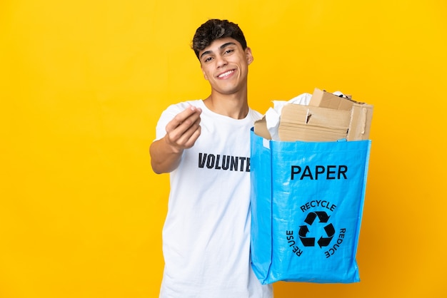 Man met een recycling zak vol papier om te recyclen over geïsoleerde gele achtergrond geld gebaar maken