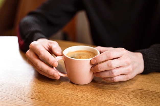 Foto man met een roze keramische kop hete zwarte koffie in zijn handen, zittend aan de houten tafel in een café.