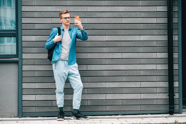 Man met een rugzak en kijkend naar smartphone terwijl hij op een achtergrond van een grijze muur staat